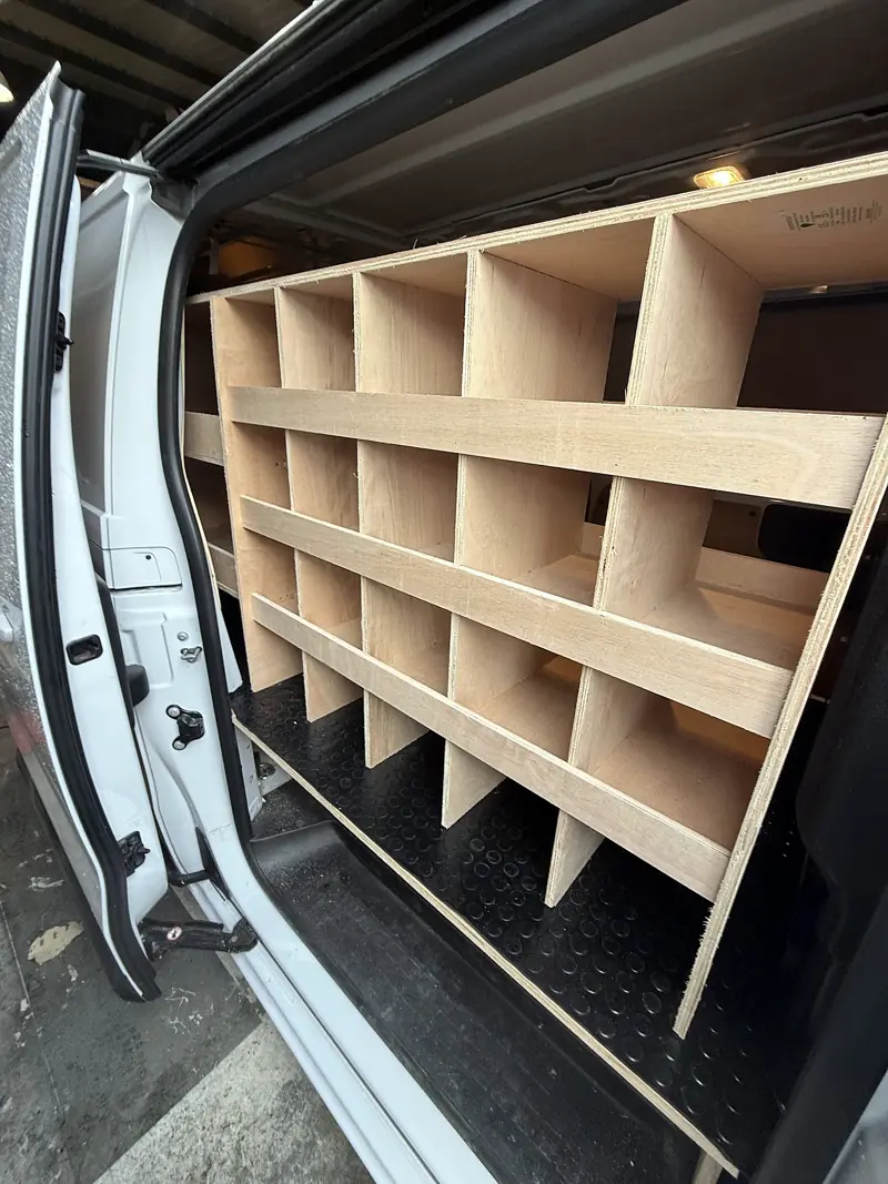 Plywood pigeonhole racking fitted down one side of a white van, seen through the open side door